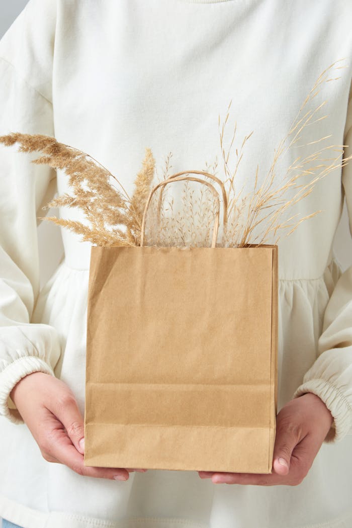 A person in a white top holds a kraft paper bag containing dry wheat stems against a white background.