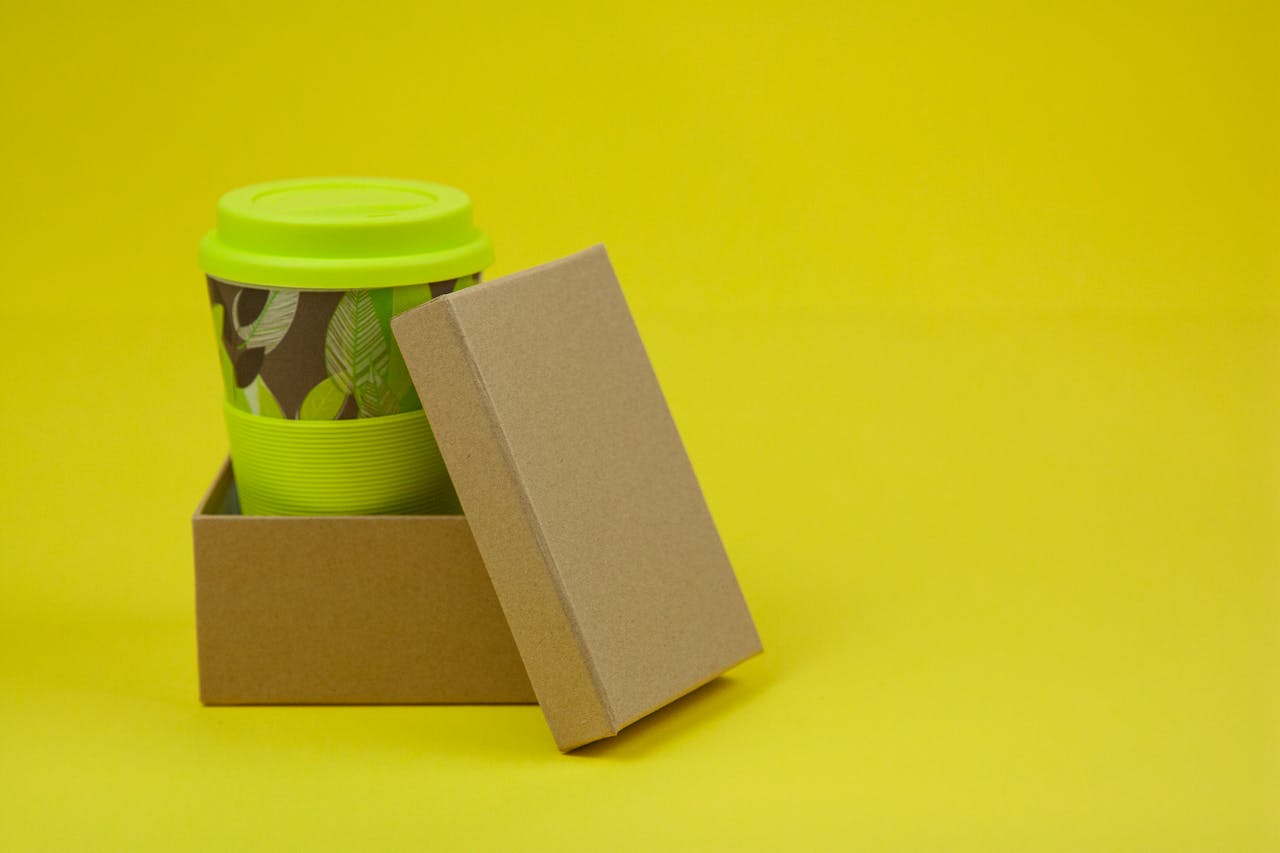 Composition of colorful paper cup with light green lid on it in small cardboard box on yellow surface